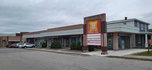 A coloured photograph of one of the car entrances of the Dynasty Centre Mall in Scarbrough showing a wide road, parked cars in front of a row of one story businesses with a large sign in red with text reading Dynasty Centre in white with a yellow stylized logo.