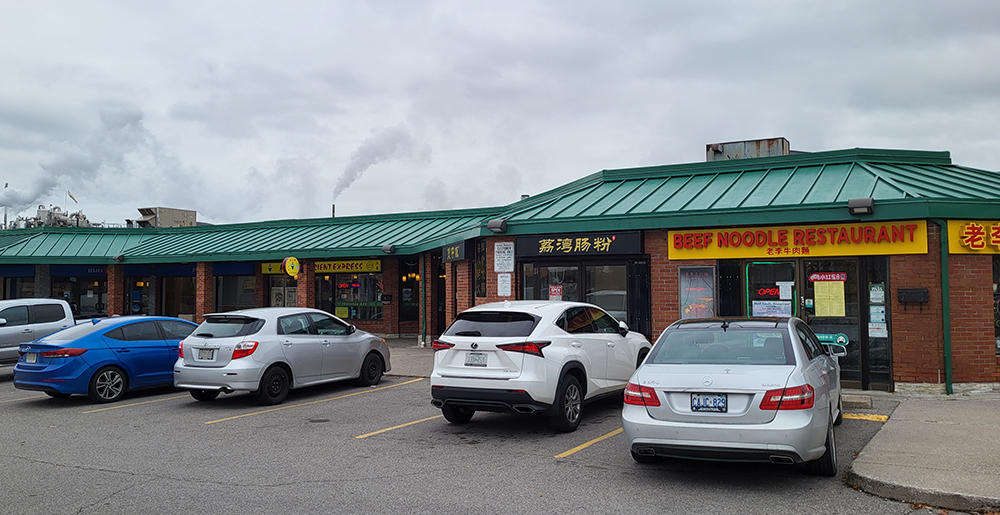 A coloured photograph of Cathay Plaza's parking lot with parked cars in front of a one story row of restaurants and businesses, including Beef Noodle Restaurant and others.