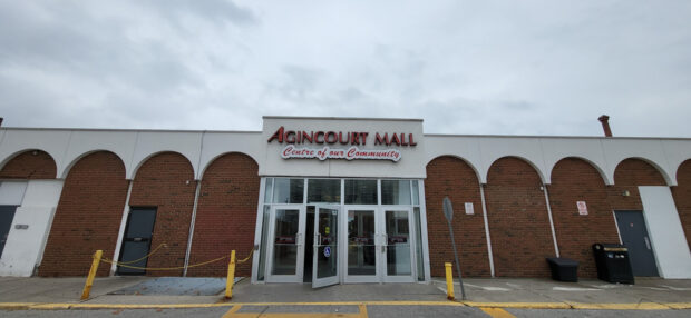 The front entrance of Agincourt Mall, written in large red letters with the subheading centre of our community against a white label, surrounded by white arches against a red brick one storey building.