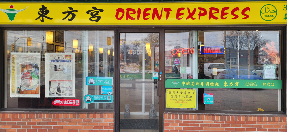 A coloured photograph of the front of the Orient Express restaurant in Torchin Plaza, Scarborough showing the restaurant's sign in yellow with red text, a window showing the inside of the restaurant and other signs in the window.