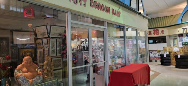 Shopfront of 'JOYS DRAGON MART' inside a mall, displaying traditional Chinese statues, art, and red decorations, with a 'For Sale' sign posted.