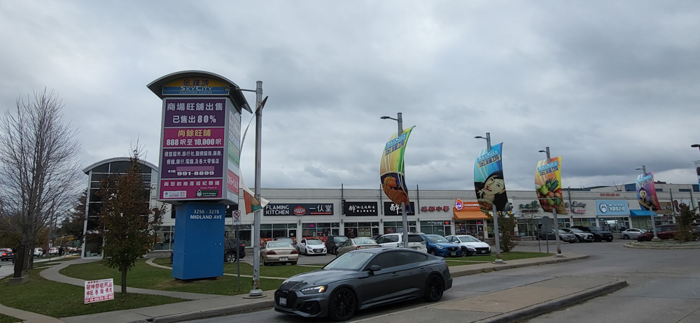 The entrance of SkyCity Mall with a car exiting past the display tower of businesses in the mall. Behind the tower is a two storey building with many storefronts amidst a busy parking lot.