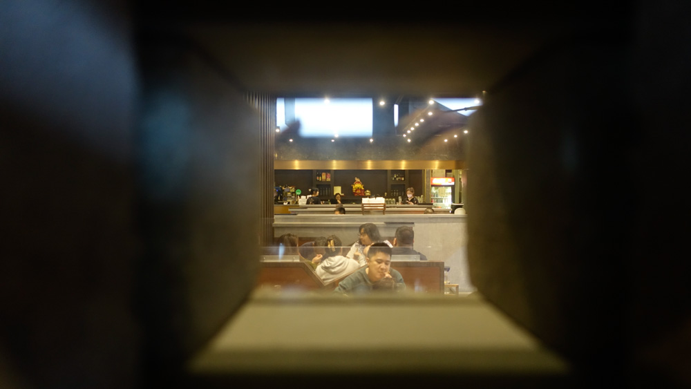 Photo taken through a square opening, looking into a brightly lit food court with people dining and a staffed counter in the background.