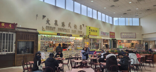 Spacious food court area with a 'Great Fountain Fast Food' sign and other eateries, with customers seated at tables under a high ceiling with skylights.