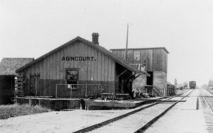 A black and white photograph of a barn-like structure with large black text reading Agincourt. The structure sits next to train tracks and depicts the one of the first railway stations in Agincourt.