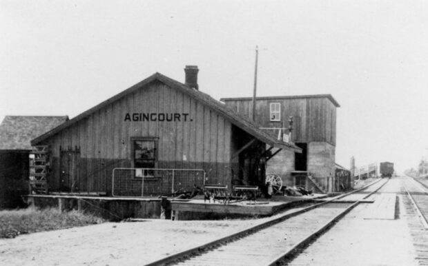 A black and white photograph of a barn-like structure with large black text reading Agincourt. The structure sits next to train tracks and depicts the one of the first railway stations in Agincourt.