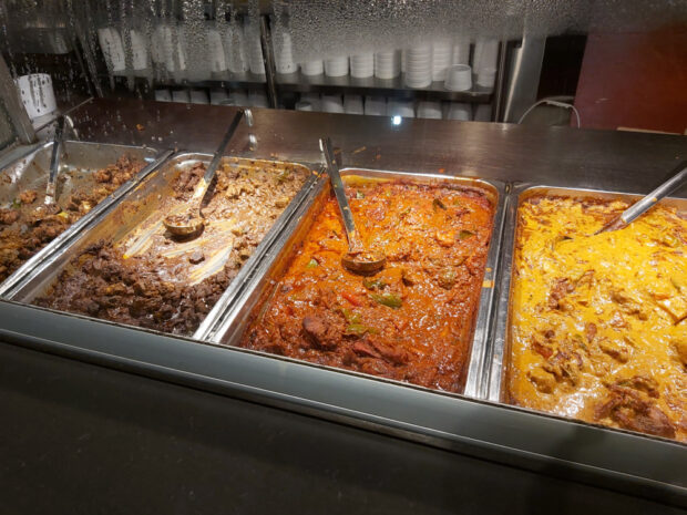 An image of three food trays at a counter, from left to right: a tray with a small amount of ground beef left, a tray with a tomato-based meat and bell pepper dish, and a tray with yellow sauce and pieces of meat.