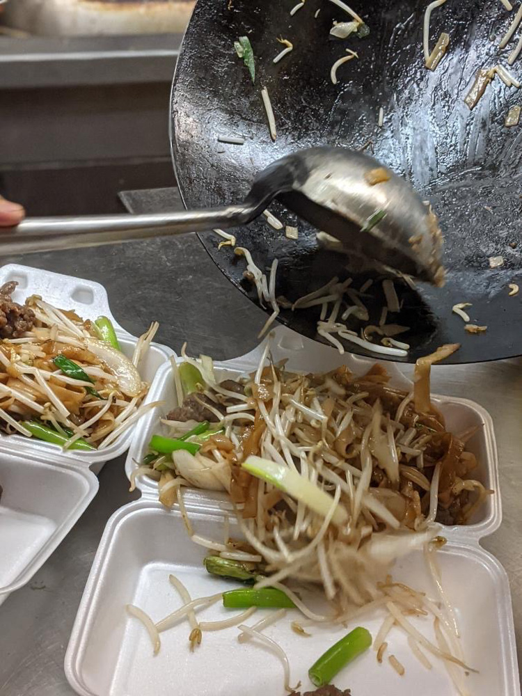 An action shot of a chef transferring a stir-fry with bean sprouts, green onions, and beef or pork strips from a wok to white foam containers.