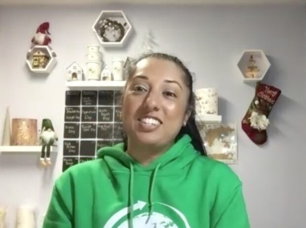 A screen capture from a video call of a smiling woman in a green sweatshirt, with a white wall and decor items behind her.