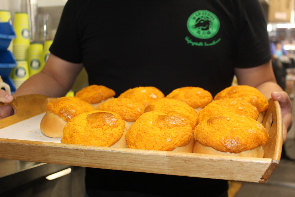 A close up of freshly baked pineapple buns that are a round bread with a bright yellow crust presented on a wooden tray, held by a person wearing a black shirt with a green logo.