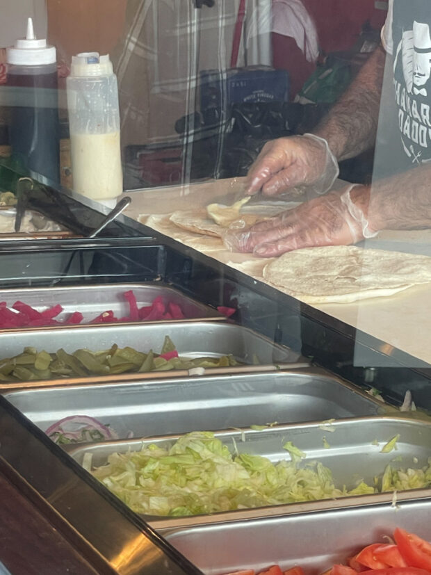 A close up image of a person preparing a shawerma wrap behind a glass partition, spreading a cream colored sauce on flatbread with trays of toppings visible in the foreground, including pickles, lettuce, and tomatoes.