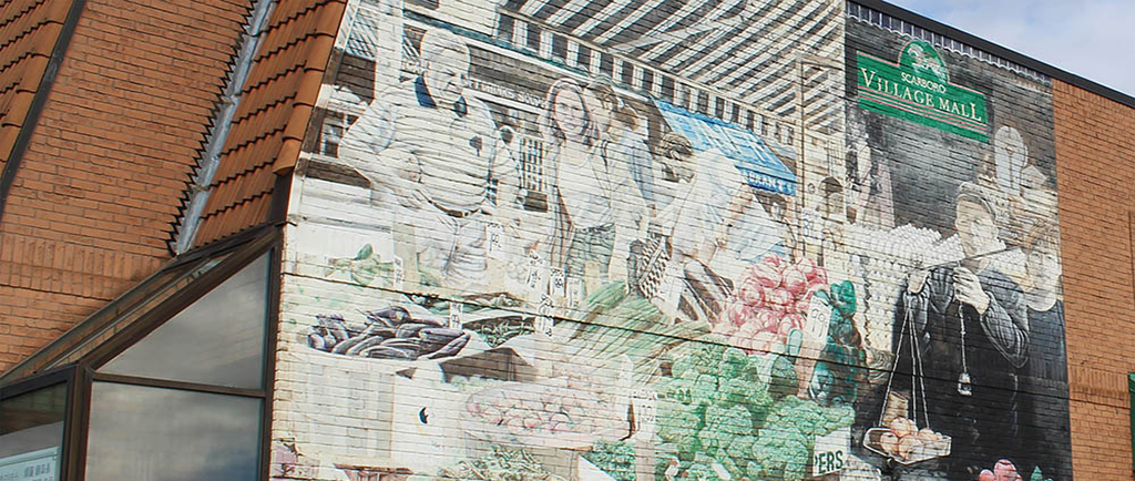 A large hand painted mural on the side of a red-bricked building featuring adult men and women shopping at a food market with a selection of vegetables laid out on a table in front of them. The market is situated under a striped veranda with a green sign reading Scarboro Village Mall in pale yellow letters.