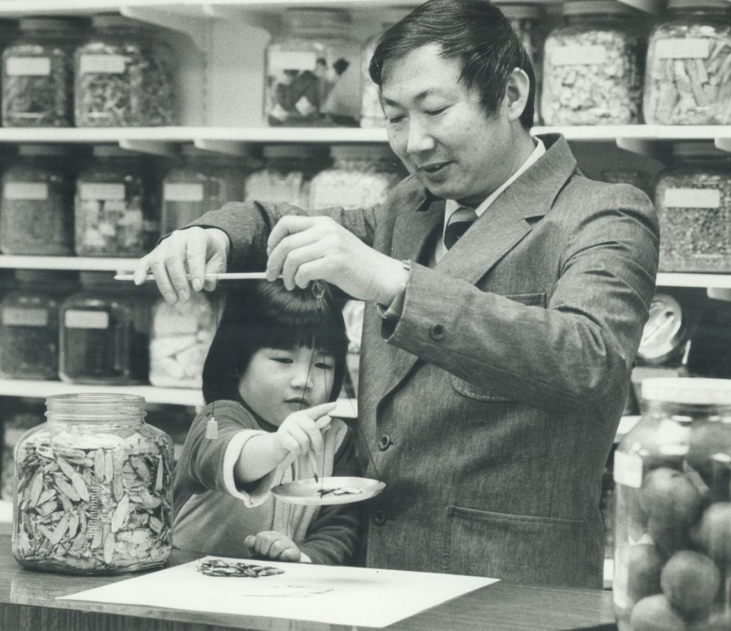 A black and white photograph of a Chinese man in a suit holding a manual scale balanced over a work surface. Next to him, a young child touching the scale with a large glass jar of organic material to the left with many shelves of similar jars containing other organic materials in a store.