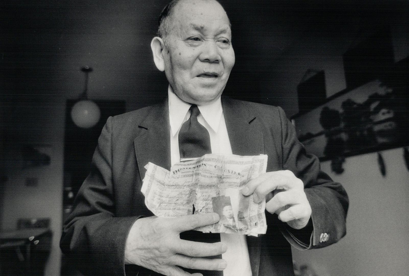 Older Chinese man in a black suit and tie holding an aged certificate with tears in the paper and writing on it, photographed in a home.