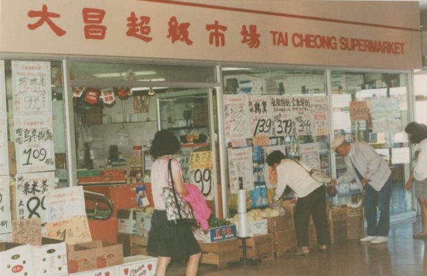 A colored film photograph of a supermarket inside a mall with large glass displays featuring multiple hand written signs with prices for goods, cardboard boxes of food items arranged on the ground in front of the glass and local shoppers selecting produce from the boxes.