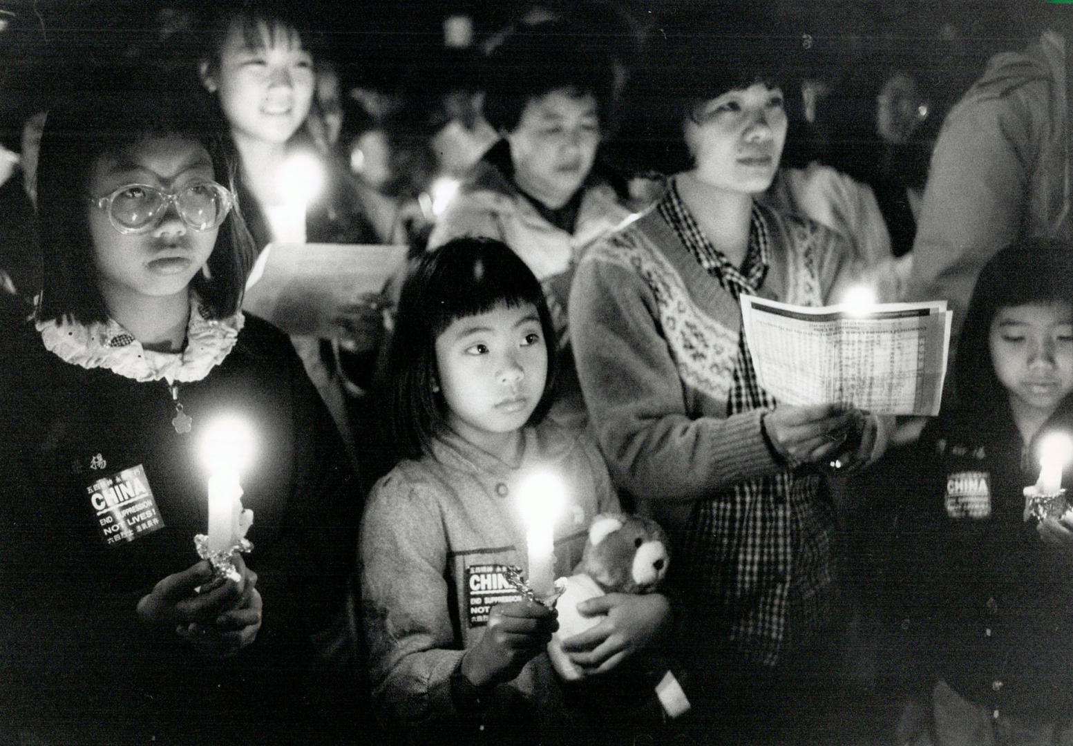 Black and white photograph of an evening gathering of Chinese women and young Chinese girls in a group holding candles, pamphlets and stuffed animals, looking sombre.