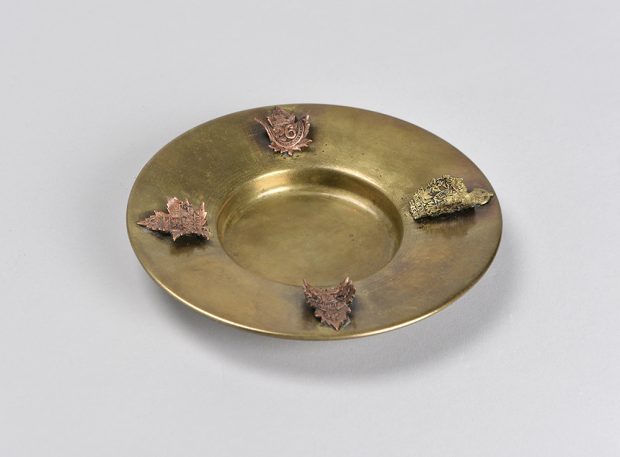 A round dulled brass ashtray. The cigarette holders a made from curved brass crests.