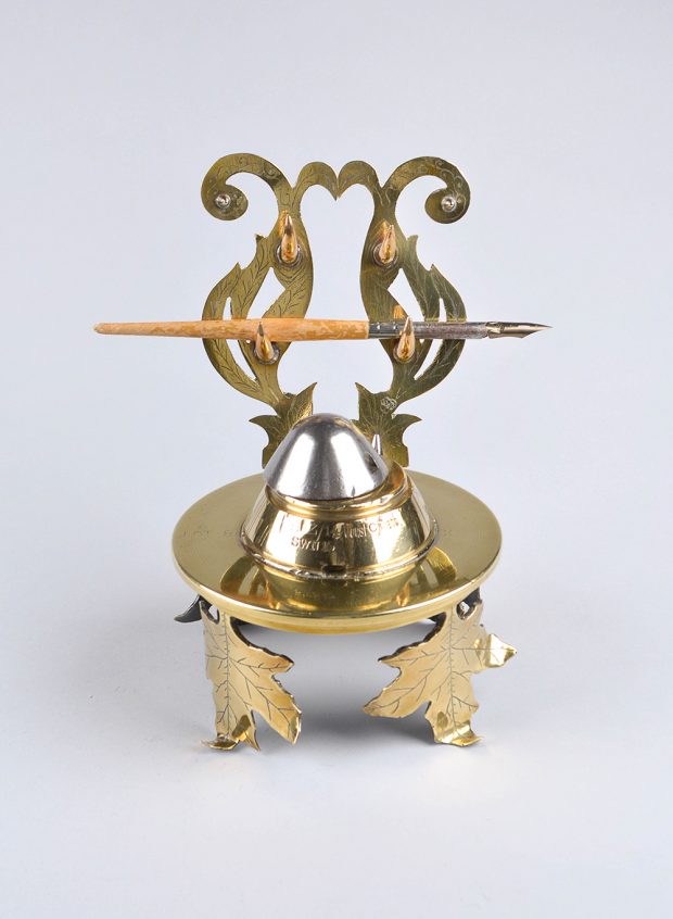 An elegant polished brass inkwell atop engraved upright maple leaves. There is a holder behind it to hold two nib pens.