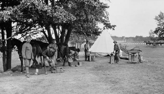 A black and white image of a portable blacksmith shop. There are several workers and horses with a tent in the ground.