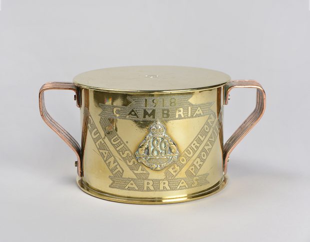 A shiny brass tobacco humidor with copper ribbed copper handles on opposite sides. There is a date and place names emerging from bands of etched background.