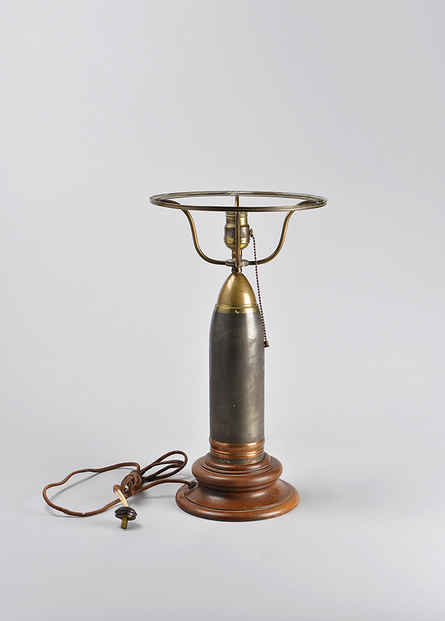 A lamp with a bevelled wooden base, the body made of a large intact bullet casing and topped with a light socket surrounded by a frame to hold a lampshade.