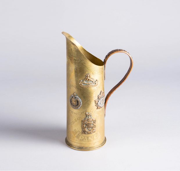 Shiny brass pitcher with an extended spout and arced handle. Covered in attached brass medals and engraved with city names.