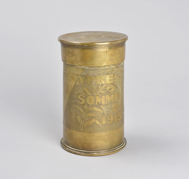 A brass humidor made from a shell casing. It is engraved with the words Ypres and some and the date 1915 over a fern leaf and background pattern.