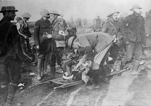 A black and white image of a chaotic war scene. In the middle, there is a soldier bent over a wounded soldier on a gurney who is clutching to his souvenir of battle.