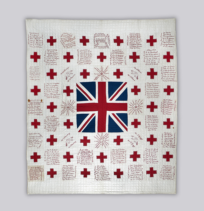 A white cotton quilt with alternating squares of red crosses and those embroidered names of people who donated money to a war cause. In the middle is a large patch of the Union Jack made of quilted material.