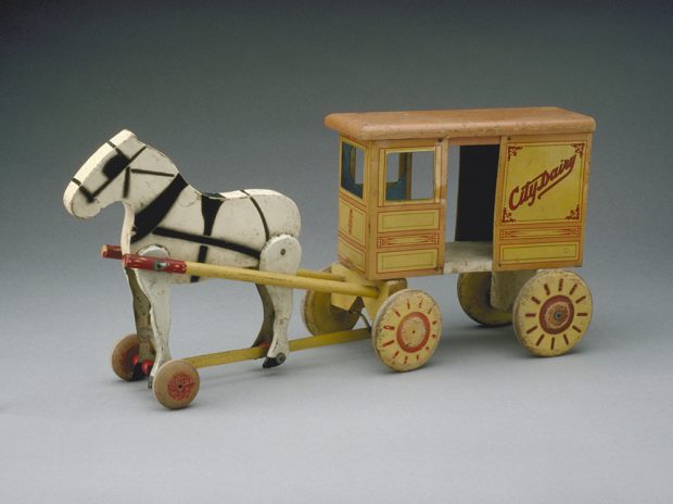 A wooden toy carving of a horse drawing a dairy carriage. The dairy carriage is painted a dull yellow, orange and red.