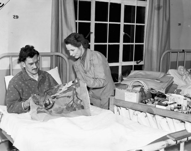 A black and white image of a Second World War soldier wearing pajamas in bed. He is doing needlepoint while being directed by a Red Cross worker.