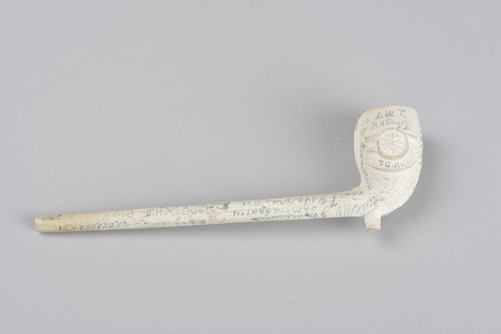 A cream-coloured pipe made from clay with embossed shapes on it. It is signed in blue ink by various soldiers.