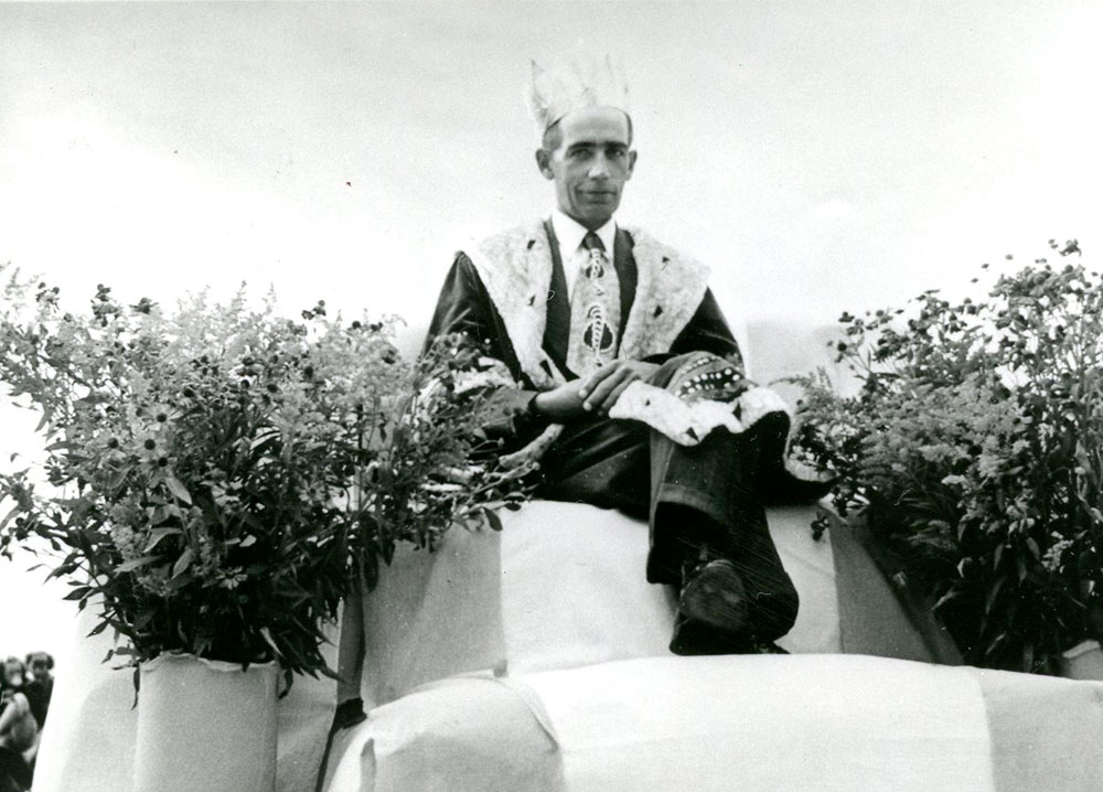 Black and white photograph of a man wearing a robe and a crown and sitting on a throne surrounded by flowers. He is the 1951 Corn King.