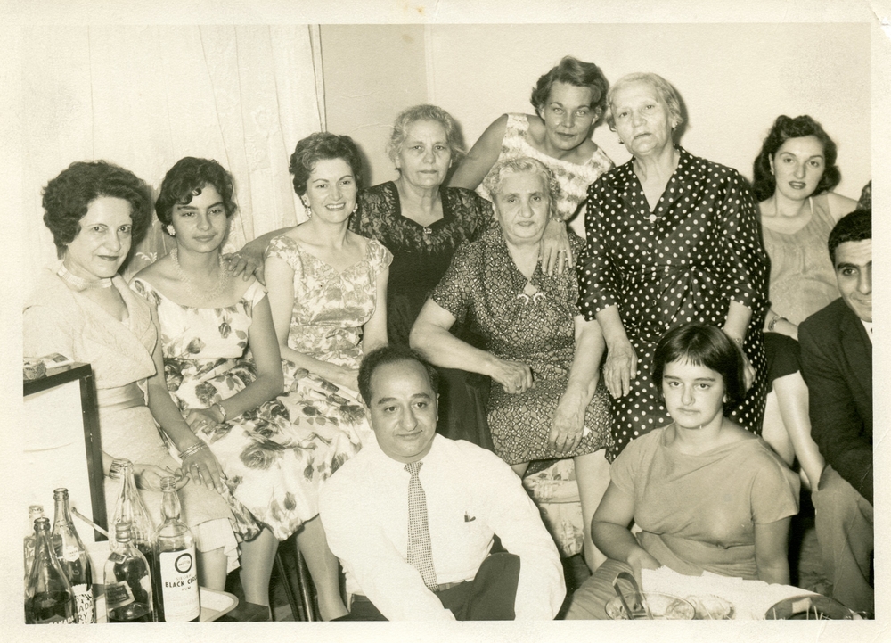 Black and white interior portrait. Nine women in dresses and two men in dress shirts or suits sit together at a gathering. Glass bottles sit on a small table in the bottom left hand corner.