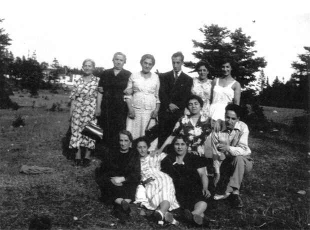 Black and white exterior portrait. Eight women and two men gather in a field with trees in the background. Five women in dresses stand in the background with one man in a suit between them; in front four women sit or kneel on the ground with one man in a dress shirt and pants.