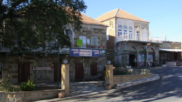 Colour streetscape. Two tan coloured stone buildings stand close together. There is a tree blocking part of the view of the left building which has a stone wall and entrance way. The right building has a patio above three large glass doors.