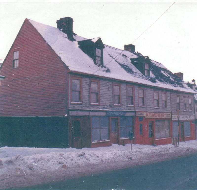 Colour streetscape of three three storey row houses. From left to right the signs above the main entrances and shop windows are City Television; A. E. Andrews; and AE Andrews. The left house is the biggest with four windows on the second floor, a dormer window on the third floor, and a chimney. The middle house has three windows on the second floor and a dormer window on the third floor. The right house has three windows on the second floor. The homes are shades of red and grey. There is another chimney between the middle and right houses.