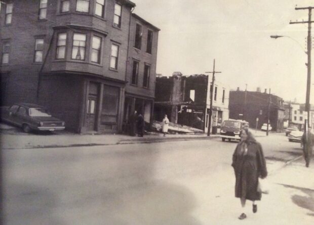 Black and white streetscape. Five people walk on the sidewalks. There are two three-storey town houses overlooking the sidewalk on the left with more two-storey row houses seen in the background.