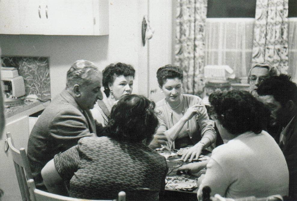 Black and white interior portrait. Five women and two men sit around a kitchen table playing cards. One of the women in the centre of the photo smokes.