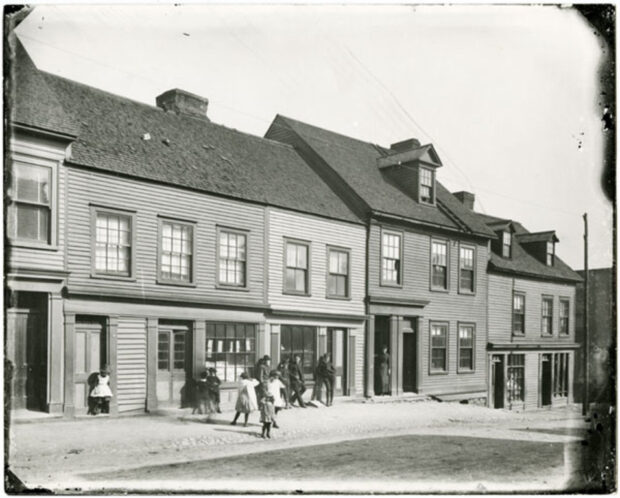 Black and white exterior image. Five townhouses in the background. Six children play in front of the homes, four men lean against one home, and one woman stands in the doorway of another home.