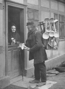 Black and white exterior portrait. A woman stands at her door holding five spoons. A man in a hat, coat and scarf holds a cane in his right hand and five wooden spoons in his left hand. He is standing on the woman’s step wearing a pack on his back filled with wooden spoons and ladles.