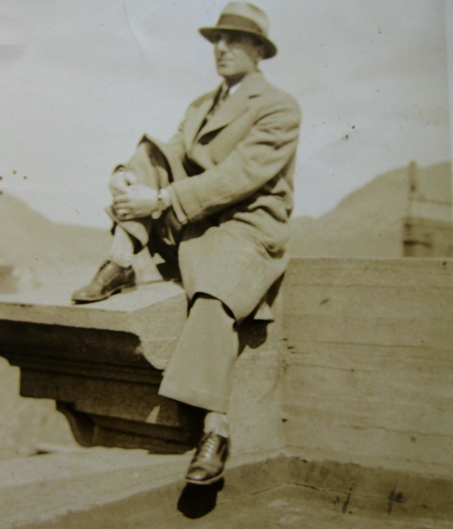 Sepia portrait. A man dressed in a fedora hat, a topcoat, pants, and dress shoes is sitting on the edge of a roof. Behind the man you can see the slope of two hills.