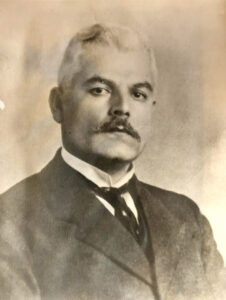 Black and white portrait of a man with light hair and a dark moustache, eyebrows and eyes wearing a suit.