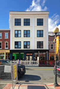 Colour streetscape. Light four-storey building in the centre reads Noah Bldg Erted 1920 Estb 1896 across the top of the building. Saltwater sign is above the main entrance. The building is flanked by two three-storey brick townhouses.