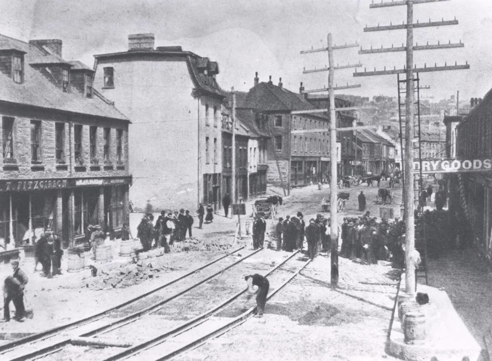 Black and white streetscape. The left hand side of the street has a mix of row and townhouses with small groups of people in front of them. Cable car tracks are being laid in the centre of the image. There is a large group of men in the right half of the photo behind two utility poles. In the background there are horse drawn carts.