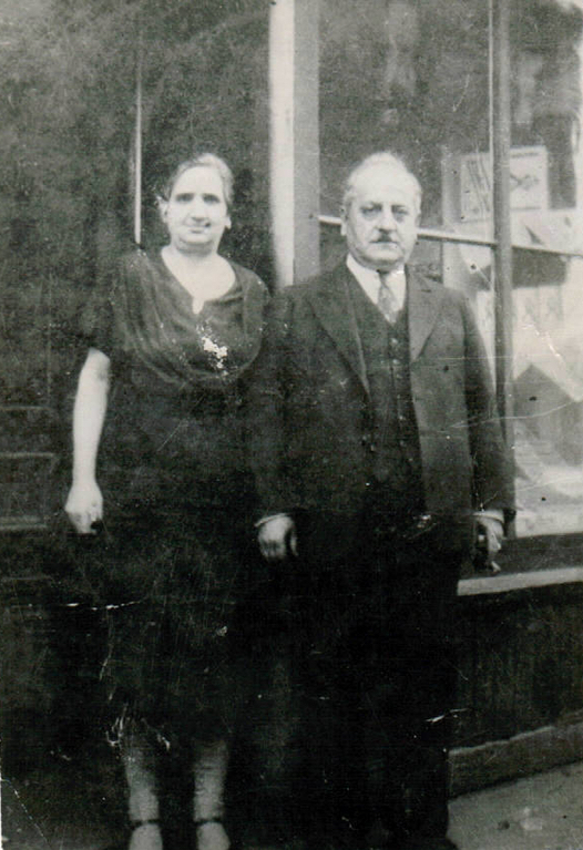 Black and white portrait. A woman in a dress, and a man in a suit stand next to each other on a doorstep. A large window can be seen behind the man.