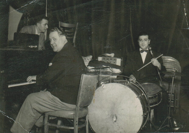 Black and white interior. Three men with musical instruments including a piano, and drum kit. The men and their instruments are in front of a large curtain.