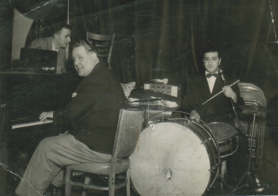 Photo d’intérieur en noir et blanc. Trois hommes avec des instruments de musique, dont un piano et une batterie. Les hommes et leurs instruments se trouvent devant un grand rideau.