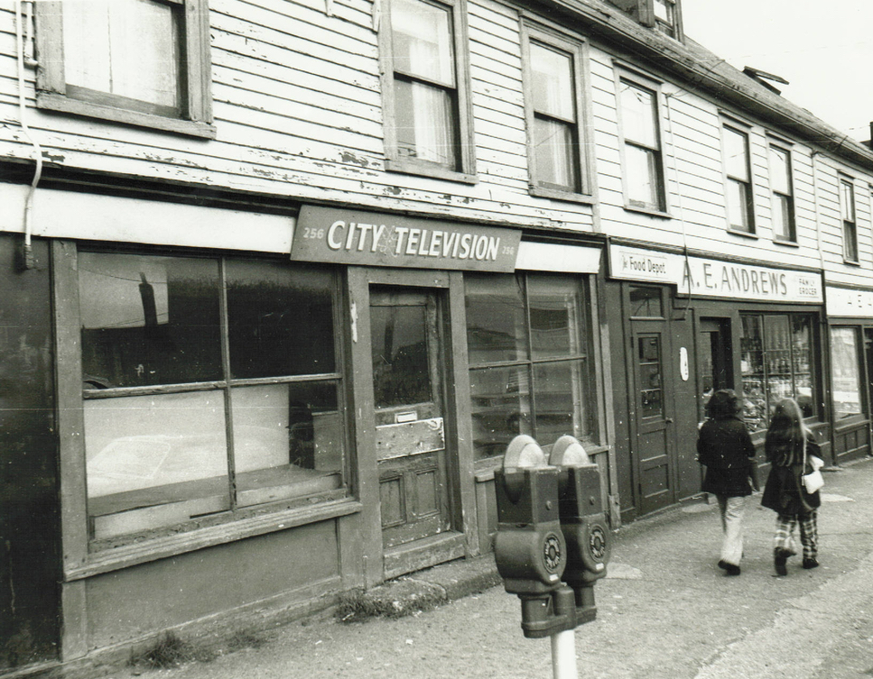 Black and white streetscape. Row houses feature City Television and A.E. Andrews signs. Two women walk down the sidewalk in front of the buildings wearing coats and pants.