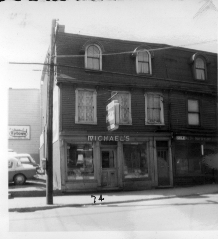 Black and white streetscape of two three storey row houses. From left to right the signs above the main entrances and shop windows are Michael's; and Sold Walsh Hairdresser. The left house has three windows on the second floor, and two dormer windows in the mansard roof on the third floor. The right house is only partially visible and has at least one window on the second floor, and one dormer window in the mansard roof on the third floor. The number 74 is handwritten on the photo in front of Michael’s. To the left of the building is a parking lot with cars and the side of a building with a sign for Uptown.